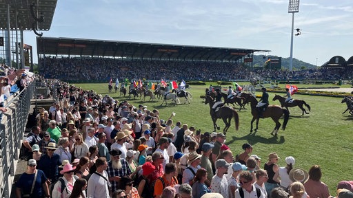 Mehrere Reiter hintereinander, die mit verschiedenen Nationalflaggen über einen Stadion-Rasen reiten.