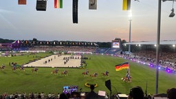 Ein Stadion, in der Mitte ein Sandfeld. Auf dem Rasen davor Reiter, einer trägt eine Deutschlandfahne.