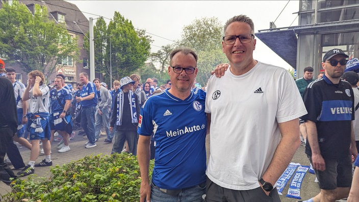 Zwei Männer stehen vor einer laufenden Meschenmenge in Fußballtrikots.