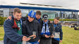 Fünf junge Männer stehen vor der Veltins-Arena und schauen auf ihr Handy.