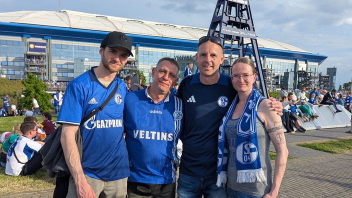 Schalke-Fan Sascha Walmsley (zweiter v. r.) steht mit seiner Familie im Arm vor dem Schalke-Stadion. Die vier tragen Schalke-Trikots.
