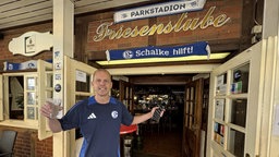 Sascha steht in einem Schalke-Trikot mit einem Glas in der einen und einem Handy in der anderen Hand vor einer Kneipentür. Er lächelt in die Kamera.