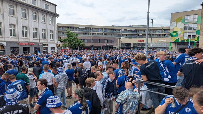 Hunderte junge und alte Schalke Fans in blauen Trikots stehen zusammen auf einer großen menschengefüllten Fläche.