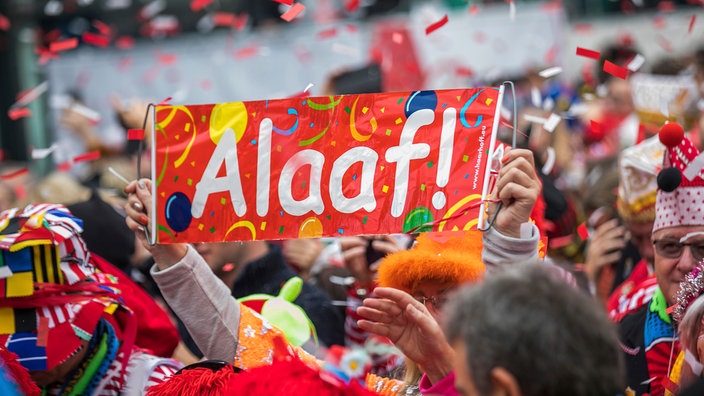 Nahaufnahme einer feiernden Menschenmenge. Jemand hälft ein Karnevals-Embleme mit der Aufschrift "Alaaf!" hoch