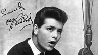 Schwarz-weiß Foto vom singenden Cliff Richard vor einem Mikrofon