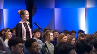 Schülerinnen und Schülern sitzen, eine Schülerin steht vor einem Mikrofon