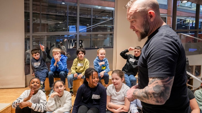 Ein Mann mit tätowiertem Arm spricht mit Schulkindern