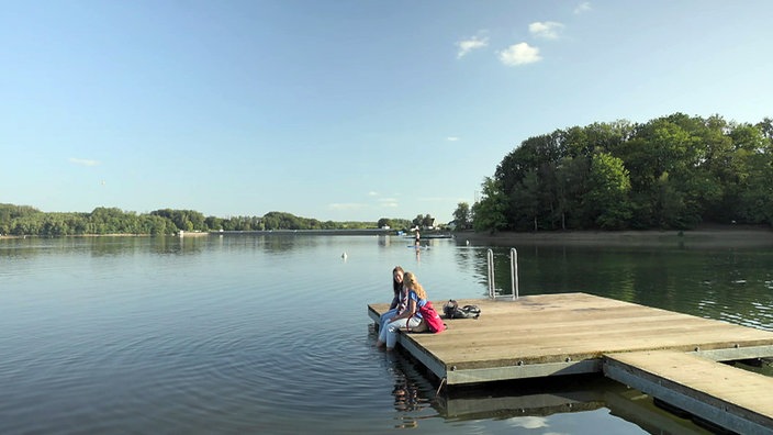Tamina Kallert sitzt mit einer Frau auf einem hölzernen Anleger auf einem Talsperrensee