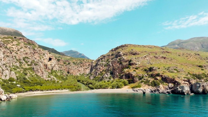 Blick vom Meer auf einen kleinen Sandstrand, dahinter ragen felsige Berge auf