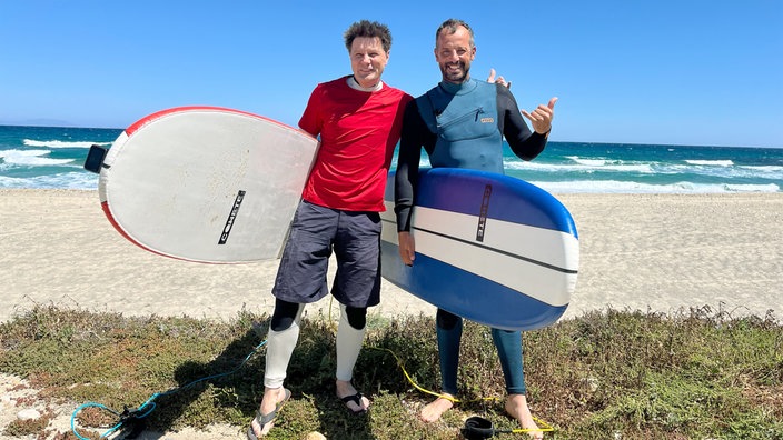 André Gatzke (l) und Manolis Orphanos mit Surfbrettern am Strand