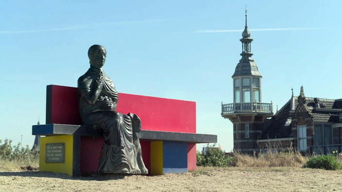 Sitzbank mit roter Rückenlehne und einer Figur, im Hintergrund der Badpaviljoen