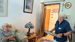 Monika Gomulia hilft einer anderen Rentnerin im Haushalt. Während Hedwig Anna Nitsch auf der Couch sitzt, läuft Monika Gomulia mit einem Stapel Wäsche durch die Wohnung.