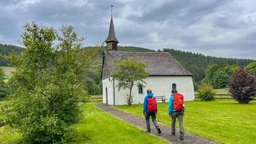 Zwei Wanderer gehen auf die Kapelle St. Agatha in Löllinghausen auf dem Wanderweg des Sauerland-Camino zu