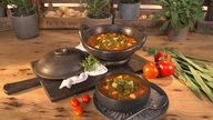 Das Bild zeigt einen Topf und eine Schüssel mit der Bohnen Suppe, präsentiert auf einem Brett mit verschiedenen Gemüse herum. 