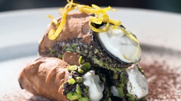 Drei Sizilianische Cannoli liegen gefüllt mit einer Ricottacreme und garniert mit Pistazien und Zitronenzesten auf einem Teller.