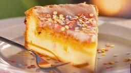 Ein Stück Kuchen mit Zuckerguss und bunten Streuseln in den eine Vanille Quarkschicht eingebacken ist auf einem lila Teller mit Kuchengabel