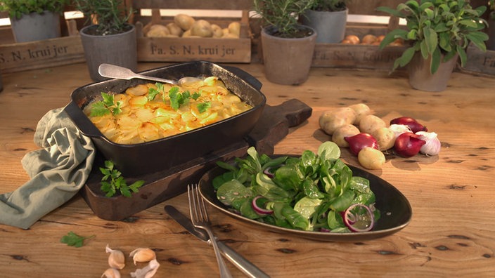 Das Bild zeigt eine auflaufform mit dem Kartoffelsalat und daneben einen flachen Teller mit dem Beilagensalat. 