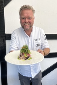Sascha Stemberg hat kurzes, graues, gelocktes Haar, ein freundliches Lächeln und trägt eine Kochuniform – auf einem Teller hält er "Sylter Krabbenbrot" in die Kamera.