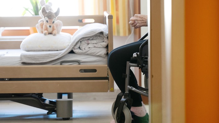 Ein Bewohner sitzt in einem Altenheim in seinem Zimmer in einem Rollstuhl. Auf dem gemachten Bett liegt ein Stofftier.