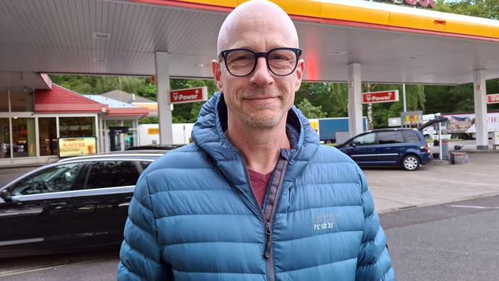Ein Mann steht lächelnd vor einer Tankstelle 