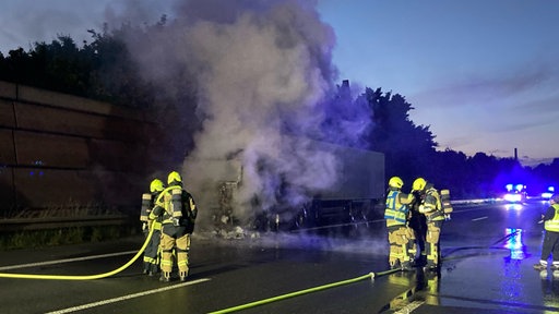 ein teils abgebrannter LKW steht auf dem Standstreifen einer Autobahn, aus dem Führerhaus steigt grauer Rauch nach oben, im Vordergrund sind Feuerwehrleute und ein Löschschlauch zu sehen
