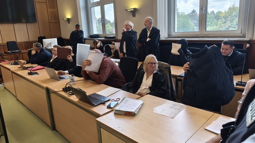 mehrere Personen im Gerichtssaal, die sich Gegenstände vor das Gesicht halten, um nicht erkannt zu werden