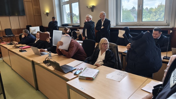 mehrere Personen im Gerichtssaal, die sich Gegenstände vor das Gesicht halten, um nicht erkannt zu werden
