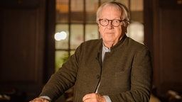 Ein älterer Mann mit Brille in einem Jacket blickt ernst in die Kamera