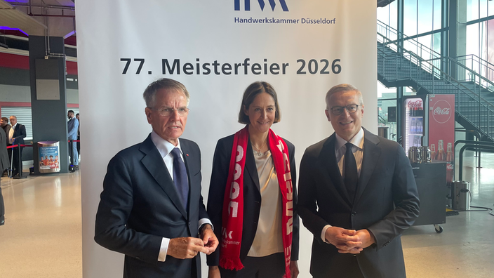 Banner zur 77. Meisterfeier 2026 mit drei Personen, die im Vordergrund stehen. Zwei Männer und eine Frau im förmliche dunkle Kleidung.