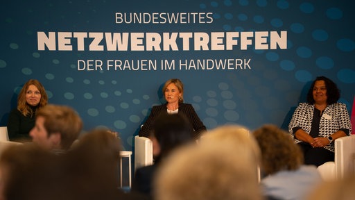 Vor einem blauen Hintergrund sitzen drei Frauen und schauen ins Publikum.
