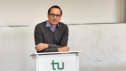 Ein Mann mit einem grauen Pullover und einem blauen Hemd darunter, der auf einem Stehpult mit einem grünen "tu" darauf vor einem Whitboard lehnt.