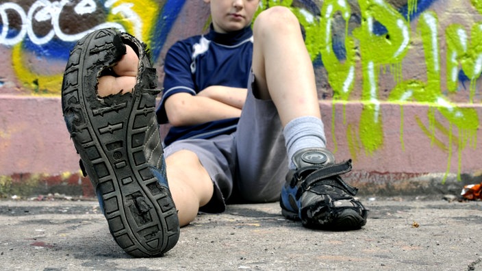 Ein Junge sitzt vor einer Wand und streckt seinen kaputten Turnschuh mit durchgelaufener, löchriger Sohle in die Kamera.
