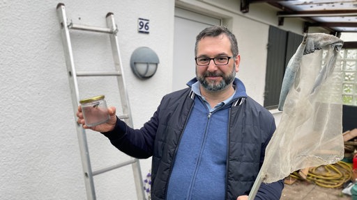 Patrick Kraus steht vor einer Gartenlaube. Er hält ein Schraubglas mit einer Asiatischen Hornisse in der einen und einen Kescher in der anderen Hand.