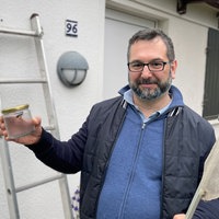 Patrick Kraus steht vor einer Gartenlaube. Er hält ein Schraubglas mit einer Asiatischen Hornisse in der einen und einen Kescher in der anderen Hand.