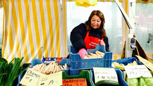 Eine Verkäuferin nimmt an einem Marktstand eine Hand voll Spargel aus einer Gemüsekiste. Rundherum steht weiteres Gemüse.