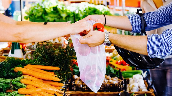 Die Arme zweier Personen vor der Kulisse eines Markstands mit verschiedenem Gemüse. Eine Person hält ein Mehrwegnetz auf, in das die andere Tomaten hineinlegt.