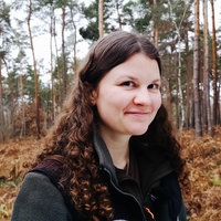 Sina Dünzer, eine junge Frau mit braunen, lockigen Haaren, steht in einem Wald und lächelt in die Kamera.