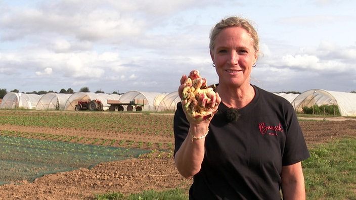 Margarete Ribbecke steht auf einem Feld. Sie schaut lächelnd in die Kamera und hält einen Ingwer in ihrer Hand.