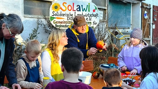 Jochen Wenzel, ein Mann mit runder Brille, brauner Kappe und gelb-blauem Kapuzenpullover, hält eine gelbe Bete in der Hand und zeigt diese einer Gruppe von Kindern.