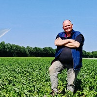 Landwirt Heinz-Georg Olligs steht auf einem grünen Feld und lächelt in die Kamera. Im Hintergrund arbeitet eine Bewässerungsmaschine.
