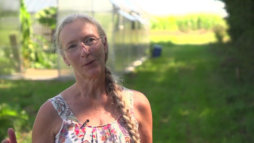 Hildegard Hendricks, eine Frau mit geflochtenem Zopf und Brille, steht inmitten von Grün, hinter ihr ist ein Gewächshaus zu sehen.
