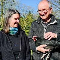 Eine Frau und ein Mann stehen nebeneinander, er hält ein schwarzes Huhn auf dem Arm