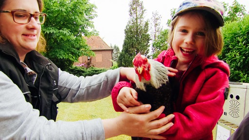 Ein Mädchen trägt ein Huhn im Arm. Neben ihr hockt eine Frau mit Brille.