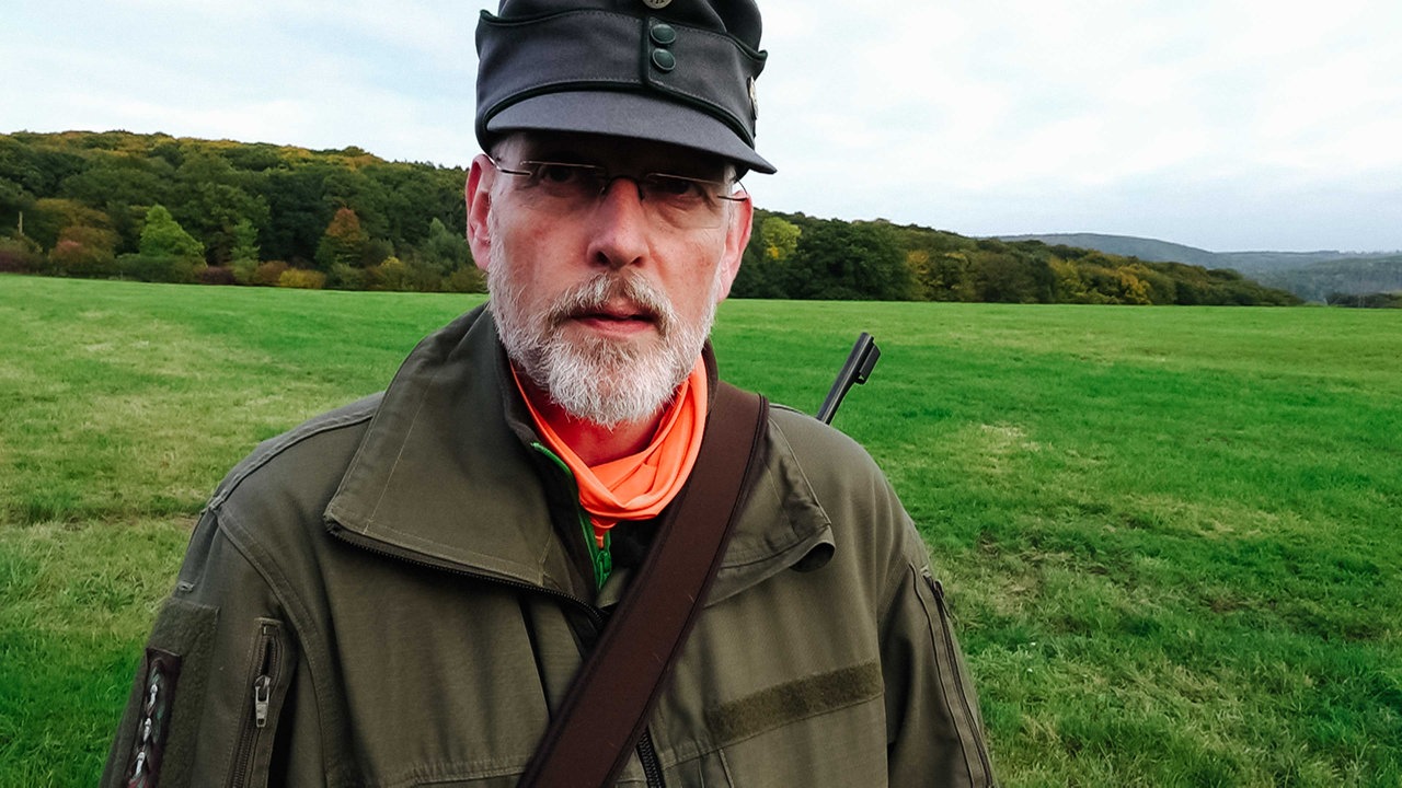 Jürgen Blasberg, silberner Bart, randlose eckige Brille, Hut auf dem Kopf, kaki-farbene Jacke, Jagdgewehr geschultert, im Hintergrund Wiese und Wald