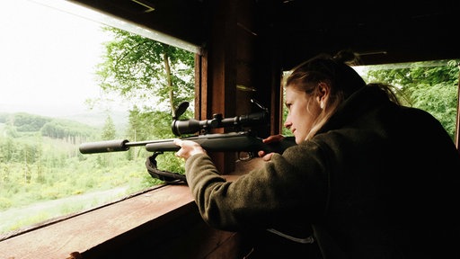 Martina Rohn steht an einer der Öffnungen ihres Hochsitzes. Mit ihrem Gewehr zielt sie in die Ferne.