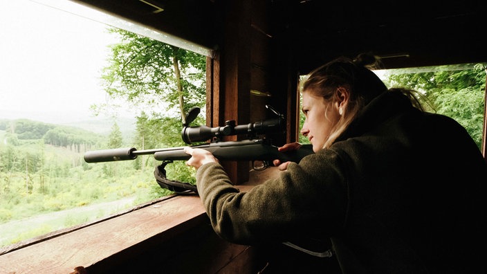 Martina Rohn steht an einer der Öffnungen ihres Hochsitzes. Mit ihrem Gewehr zielt sie in die Ferne.