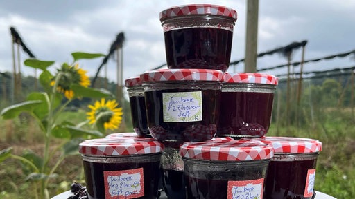 Auf einem Tisch sind mit Junibeeren-Marmelade gefüllte Marmeladen-Gläser mit weiß-roten Deckeln gestapelt
