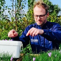 Ein Mann mit braunen Haaren, Brille und blaunem Hemb auf einer Wiese. Vor ihm ein weißer Eimer mit lila Blüten drin