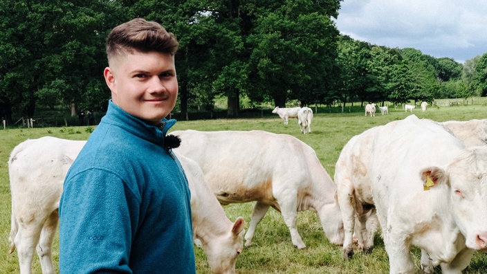 Hannes Knickenberg steht auf einer Wiese, hinter ihm weiße Kühe. Er lächelt.