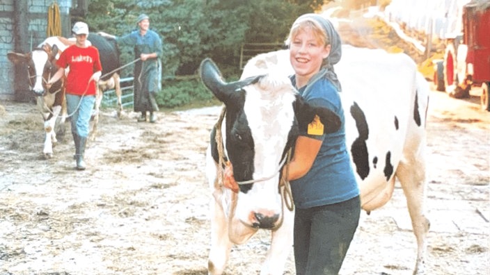 Ein älteres Foto der jungen Katrin Engels, die auf dem Hof ihrer Familie steht und lächelnd den Hals einer Kuh umarmt.